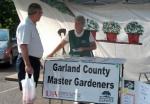 Garland County Master Gardener talking with man at exhibit tent in parking lot plants and sign