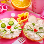 bagel with cottage cheese toped with chives and veggies arranged to resemble a cat face and mouse face on the other slice