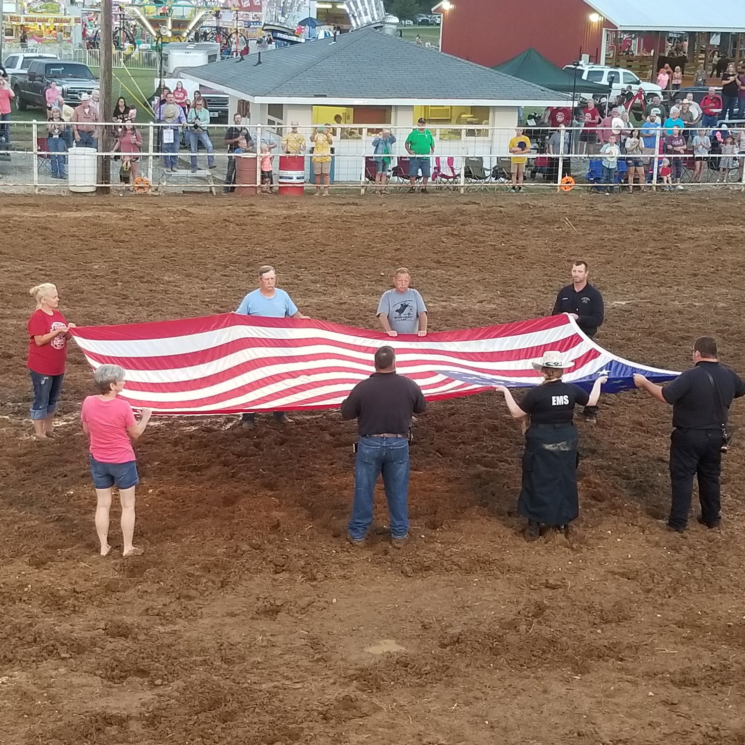 8 people folding a US flag on a dirt floor in front of a crowd