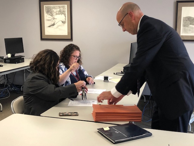 Two women sitting at a table review paperwork from a man who is standing