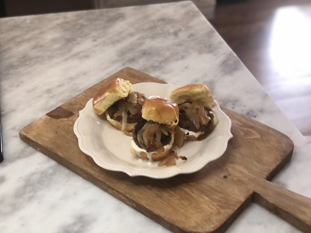 Three deer sliders with caramelized onions and aioli on a white plate placed on a wood cutting board. 