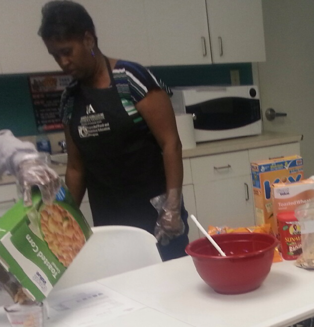 Drew County EFENEP Program Assistant sharing food demonstration, Female in apron standing behind table with food on it 