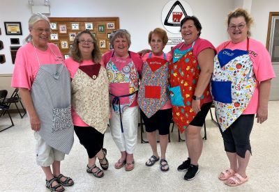 Six smiling EHC ladies standing in a row modeling their colorful sample aprons they made for a 4-H youth summer sewing camp.