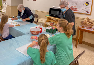 Three girls sitting at a table with two volunteer ladies standing and showing them how to dip pretzels in melted chocolate.