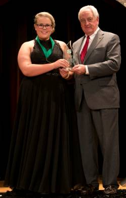 Girl with glasses wearing long black dress shaking hands and receiving award from man wearing gray suit, white shirt and red tie.