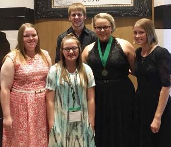 Girl in coral print dress, girl wearing glasses and aqua dress with black tie dye stripes, girl wearing glasses and black dress and medal hanging from green ribbon, girl with chin-length hair wearing black dress, boy standing behind the girls and wearing a black shirt