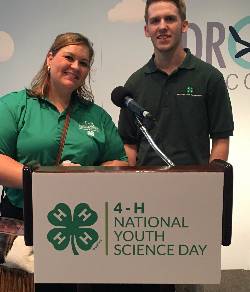Woman wearing green shirt with 4-H logo and teenage boy wearing green shirt with 4-H logo standing behind a podium with a white sign hanging from the front of it. A large 4-H clover is on the left side and the words, "4-H National Youth Science Day" on the right side.