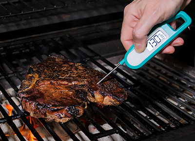 person inserting a meat thermometer into a steak on a grill