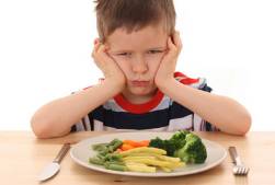 Young white boy wearing red, white, and blue striped shirt, with his elbows on the table and his head resting in his hands which are on either side of his head. Unhappy look on his face. A white plate with green beans, orange carrots, green broccoli, and celery strips. A silver knife is on the left side of the plate and a silver fork on the right side of the plate