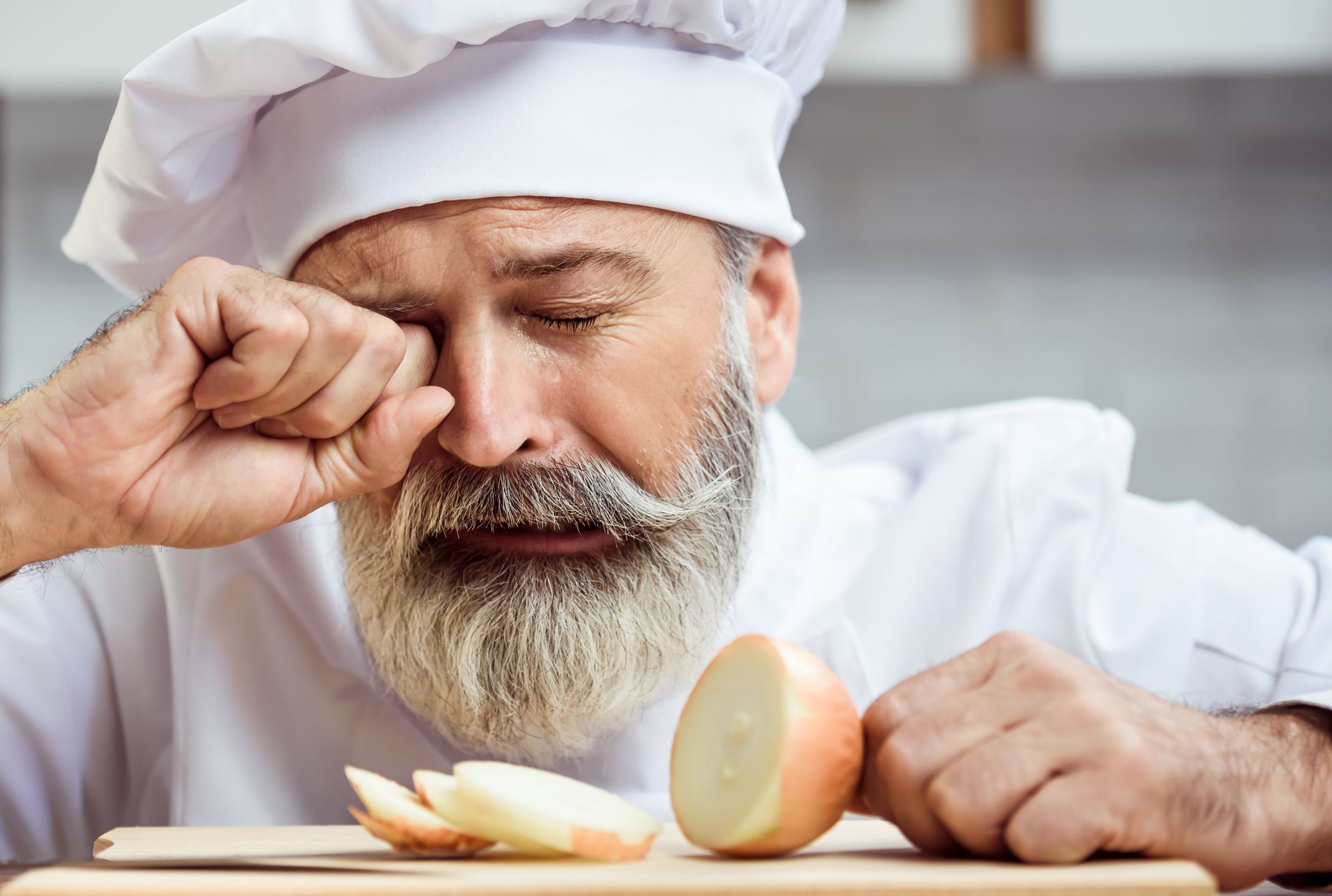 man in chef outfit standing over cut onions wiping tears away