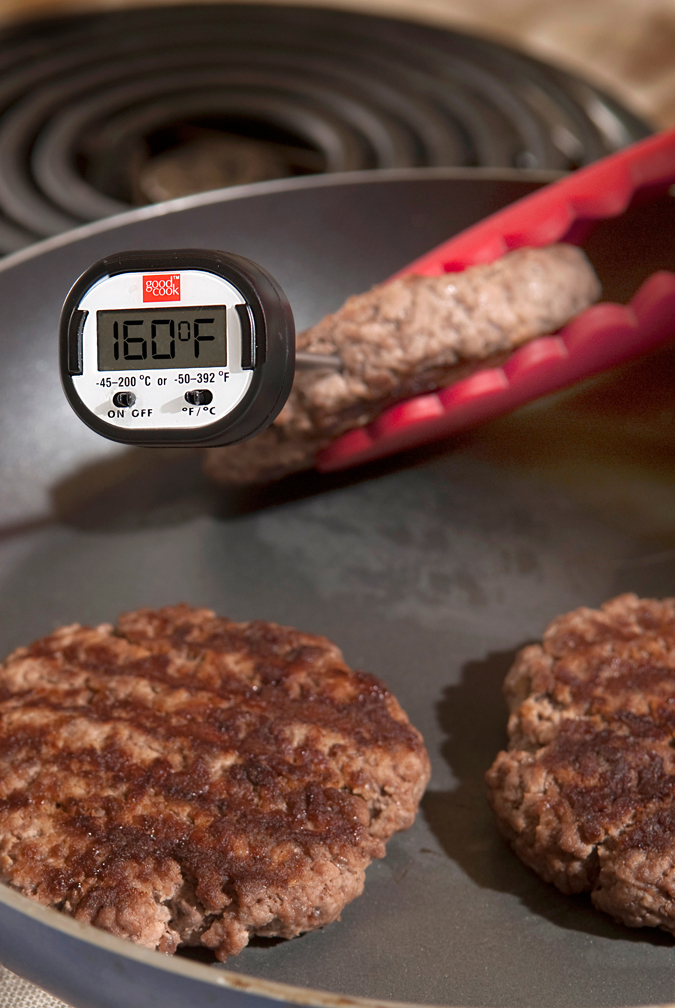 hamburger patty on the grill held between tongs with thermometer showing 160 degrees F inserted in center of meat.
