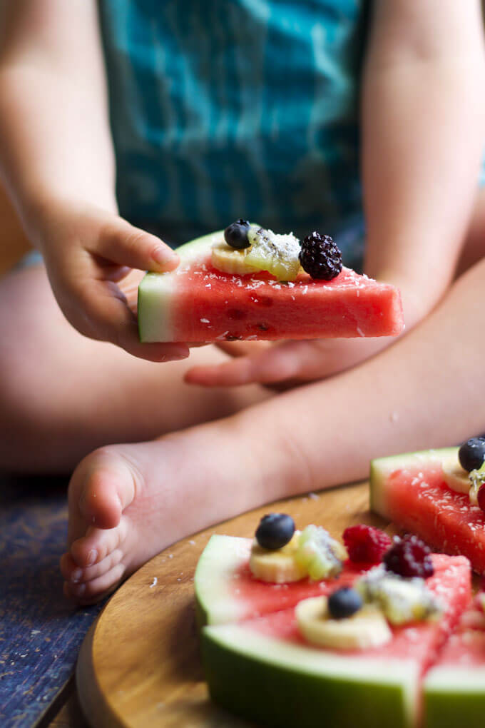 child sitting with watermelon sliced in a circle on a wooden tray with berries and bananas on top of sliced watermelon