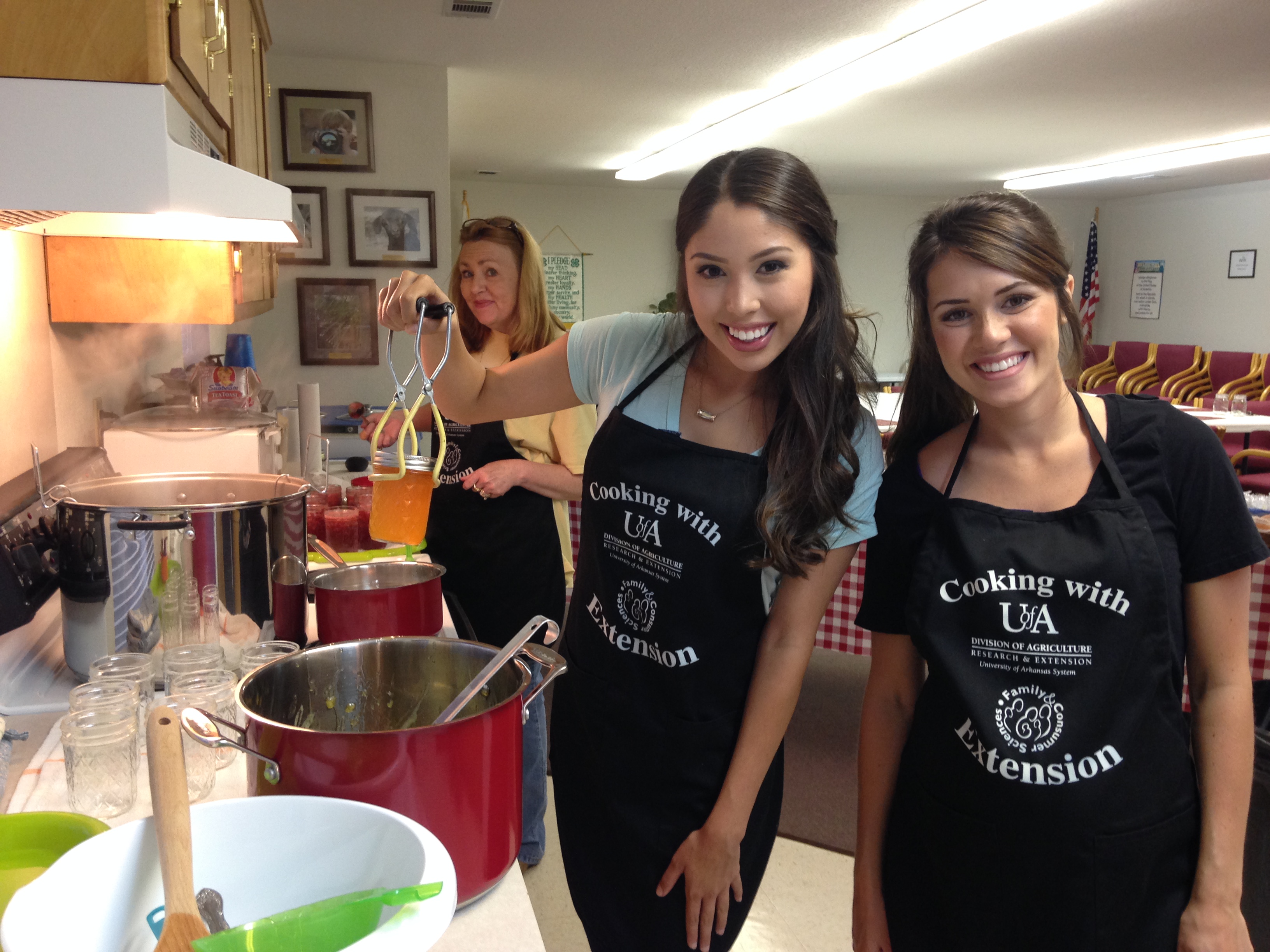 Two women holding a freshly canned jar of jelly while a third woman smiles in the background