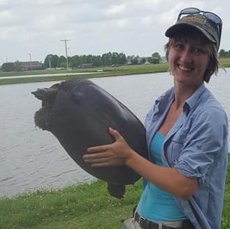 A young lady in a blue shirt and cap holding a turtle with a pond in the background