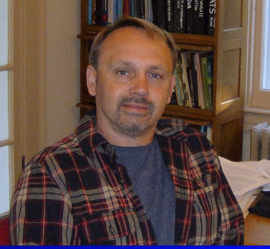 A man in a plaid shirt sitting in an office with books in the background