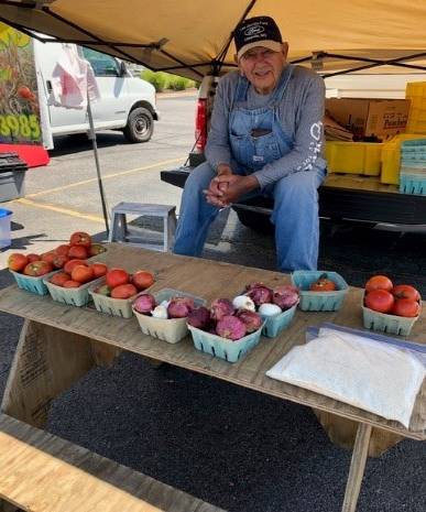 Photo of a man sitting behind a table with an assortment of vegetables in cartons for sale