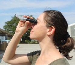 A woman holding a handheld refractometer in the air and looking into it