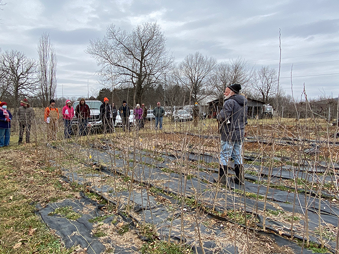 Guy Ames standing in a few rows of saplings speaking in front of a crowd on his farm