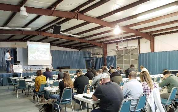 Man standing on a stage giving a presentation to a group of people sitting at tables taking notes