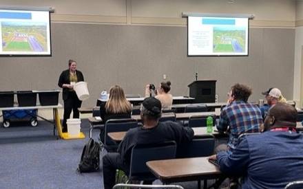 Presentation in a conference room with people sitting at tables. Speaker is holding a bucket.