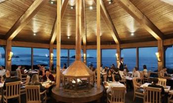 People dining out sitting at tables in a restaurant with the ocean outside the windows that is the wall to the restaurant.