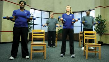 Two women in front row and two men in back row standing by their chairs holding weights in both hands palms up at waist level.