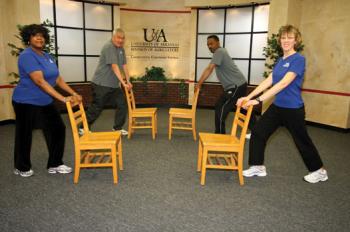 Two women in front row and two men in back row holding onto the back of a chair while doing a calf stretch.