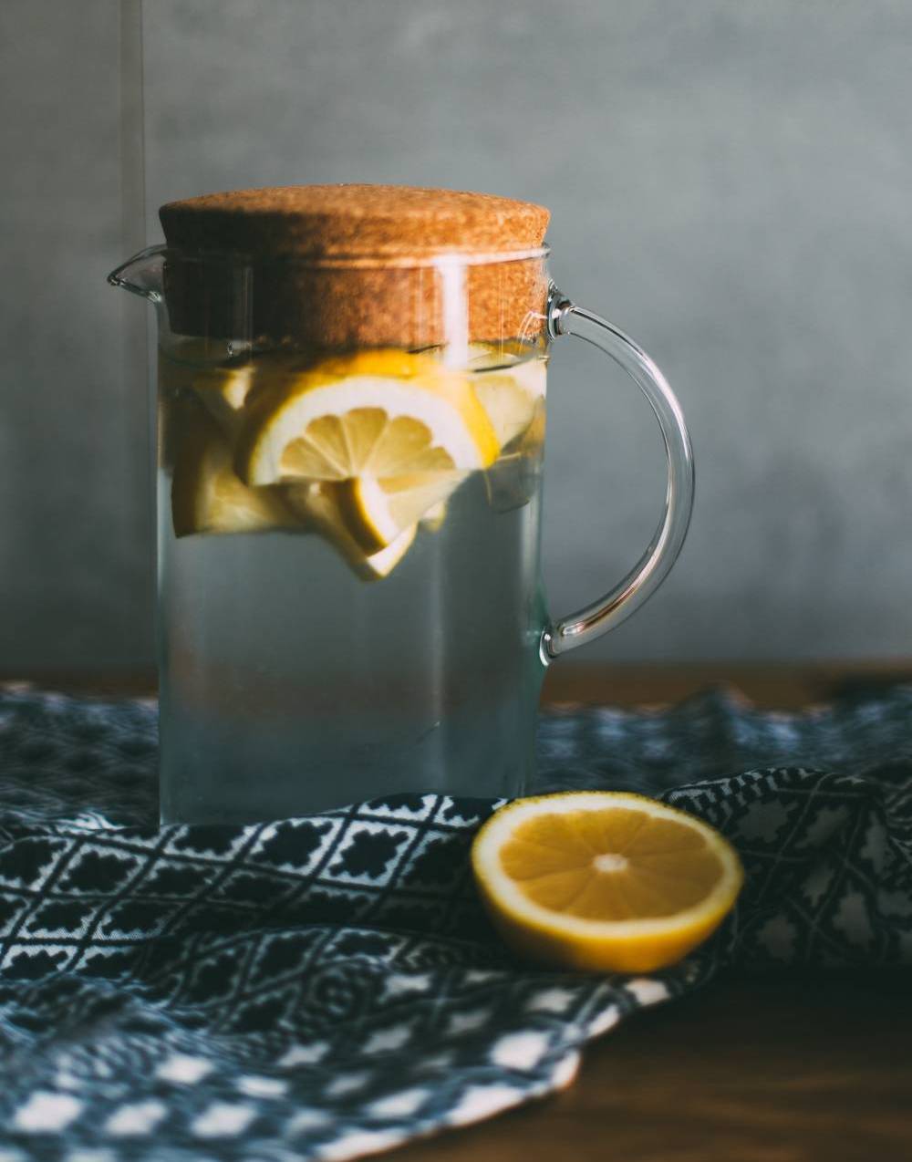 Clear pitcher on a table with water and sliced lemon in it. A half lemon sits at the base of the pitcher.