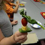 A student creates a healthy snack on a stick with fresh spinach leaves, a cube of cheese, and a cherry tomato