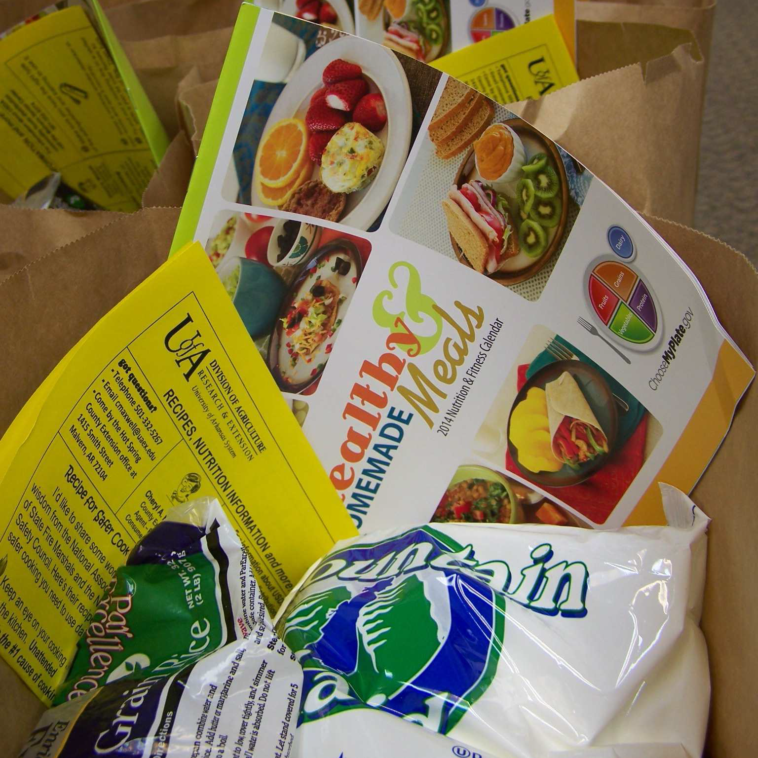 Paper grocery bag full of commodities such as rice and dried beans. Also includes a monthly calendar of recipes and a yellow newsletter telling how to use the commodities.