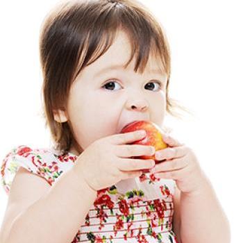 Preschool girl in a pink dress with red flowers takes a bite of a red and yellow apple