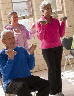 Adults enjoying light weightlifting in a bright and airy community center recreational room. One older gentleman is seated while two ladies stand alongside him.