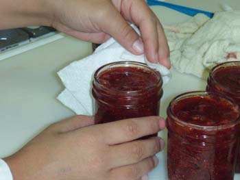 Three small jars of fruit sitting on a counter with hands holding a napkin wiping off one of the jars preparing it for the lid.