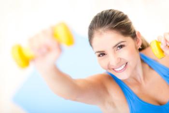 Woman wearing blue top is smiling at the camera while holding a hand weight with her right arm stretched above her head and the weight in her left hand is at her shoulder.