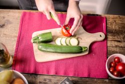 Hands slicing a cucumber on a wooden cutting board.